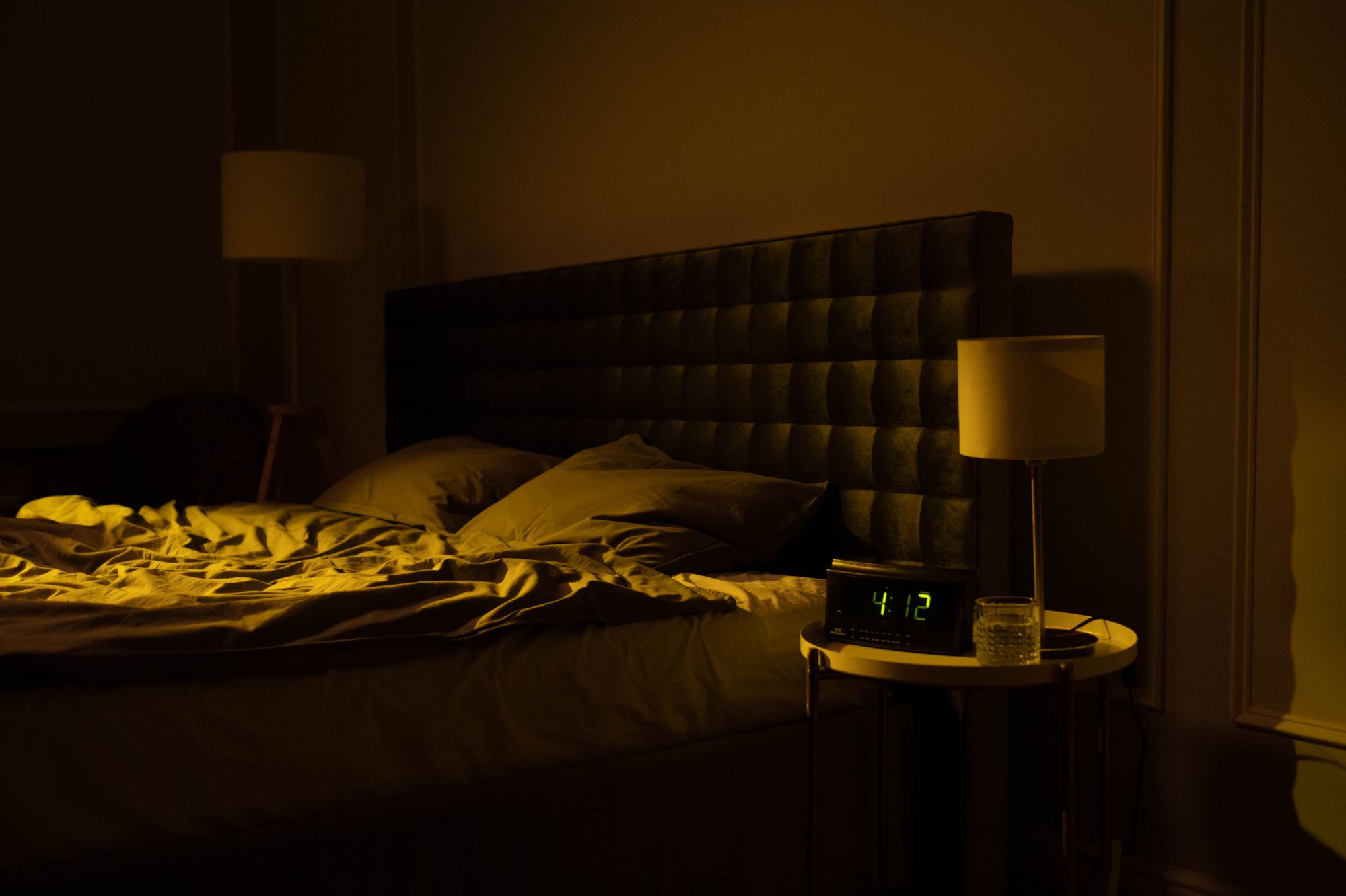 A dark bedroom lit only by a warm bedside lamp, a clock on the nightstand reading 4:12