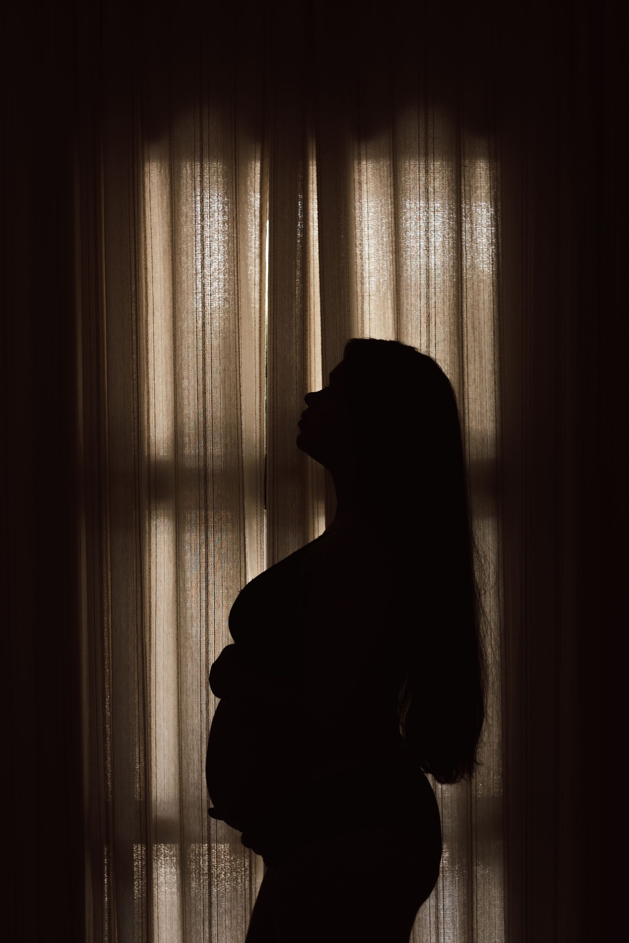 A pregnant woman standing at a curtained window in low light, looking up, one hand resting under her belly